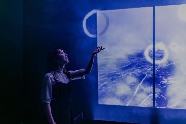 A side view of a young woman, who is smiling and reaching up with one arm to touch a floating circle, which looks like it is made of white smoke. Behind her is an image of rippling water and more floating circles.