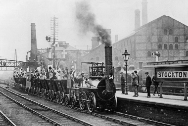 An old black and white photos of a steam train is coming into a station. There are bridges and chimney stacks in the distance. The train is a replica of Locomotion No. 1 with a large funnel with dark smoking coming out top. 