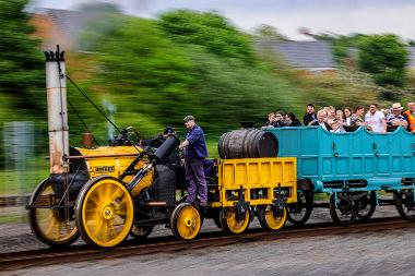 A replica Locomotion No.1 locomotive in bright yellow is in motion. The background is blurred to show the train is in motion. There is a driver stood on the plate to the side of the train. The turquoise carriage behind has people in it.